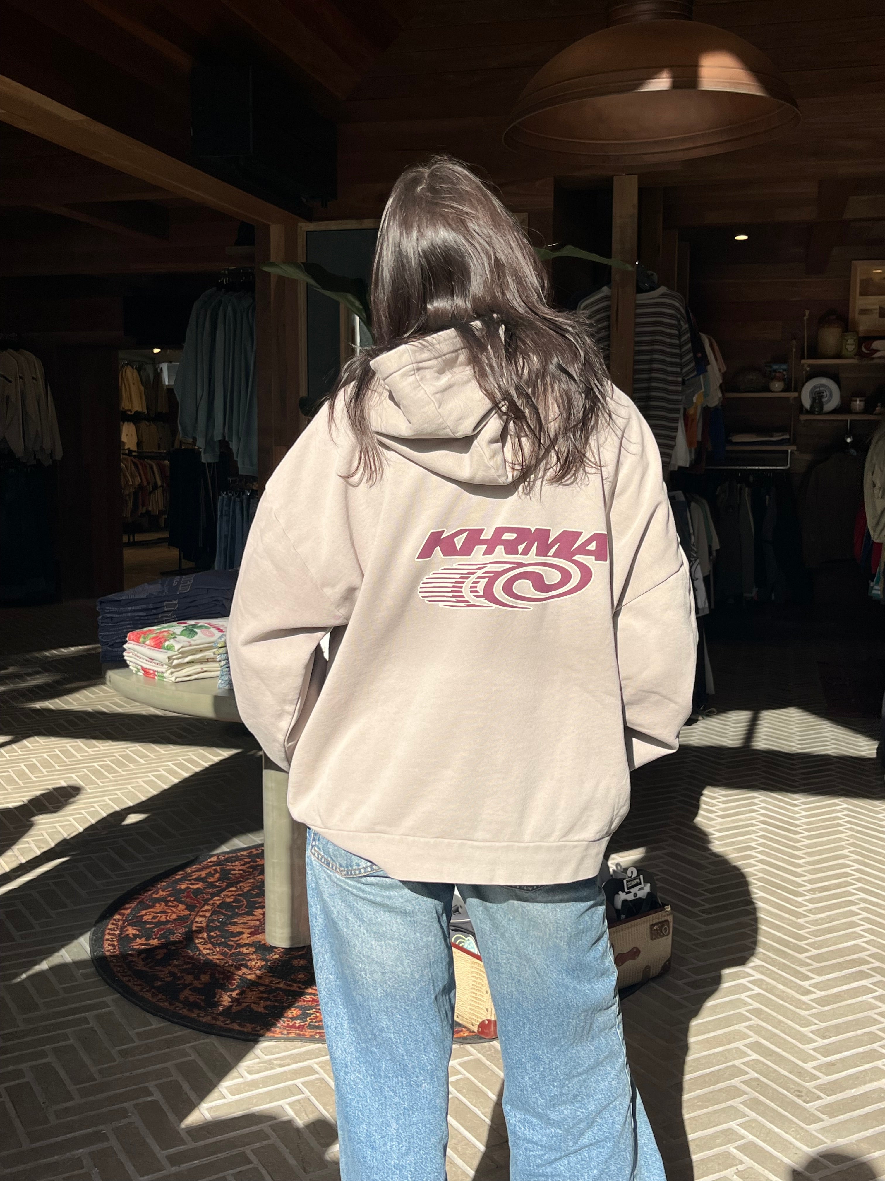 Person wearing a beige hoodie with a logo, standing indoors on a patterned floor.