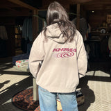 Person wearing a beige hoodie with a logo, standing indoors on a patterned floor.